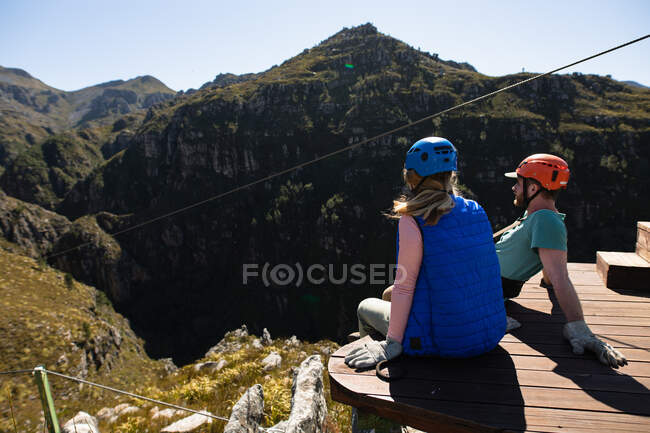 Vista lateral de la pareja caucásica disfrutando del tiempo en la naturaleza juntos, con equipo de forro de cremallera, sentado en la cubierta en un día soleado en las montañas - foto de stock