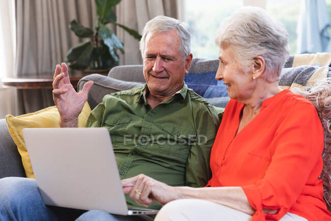 Una pareja de ancianos jubilados caucásicos en casa sentados en un sofá en su sala de estar, hablando y sonriendo, utilizando un ordenador portátil juntos, pareja aislante durante coronavirus covid19 pandemia - foto de stock