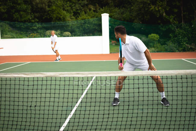 Un Caucasien et un homme de race mixte portant des blancs de tennis passant du temps sur un court ensemble, jouant au tennis par une journée ensoleillée, tenant des raquettes de tennis — Photo de stock