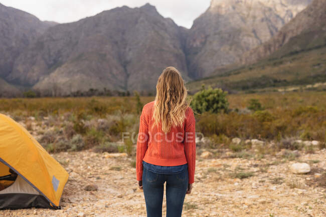 Vista posteriore di una donna caucasica che fa un buon viaggio in montagna, in piedi vicino a una tenda, guardando le montagne — Foto stock