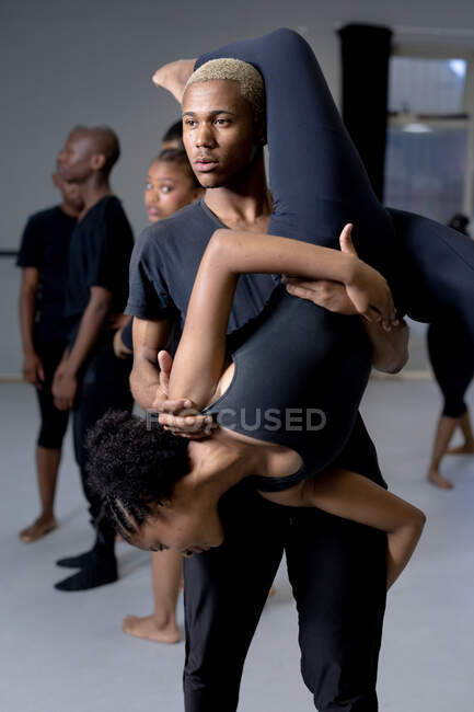 Vista frontale da vicino di una razza mista adatta a ballerini moderni di sesso maschile e femminile che indossano abiti neri che praticano una routine di danza durante una lezione di danza in uno studio luminoso, l'uomo sta tenendo la donna in posa a testa in giù — Foto stock