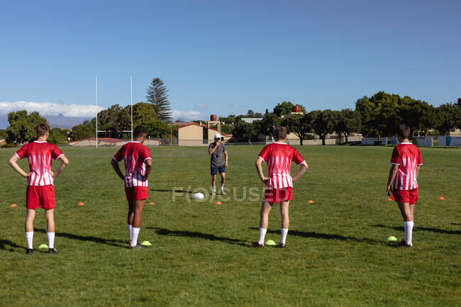 Rückansicht von vier multiethnischen männlichen Rugby-Spielern, die ihre Mannschaftskleidung tragen, auf dem Spielfeld stehen und den Anweisungen ihres Trainers lauschen — Stockfoto