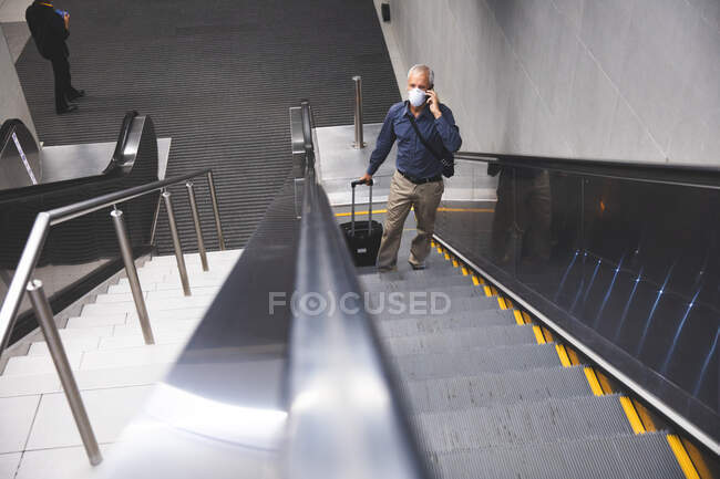 Homme caucasien âgé, portant un masque facial contre le coronavirus, covide 19, utilisant un escalator dans une station de métro, parlant sur son smartphone, et tirant une valise. — Photo de stock