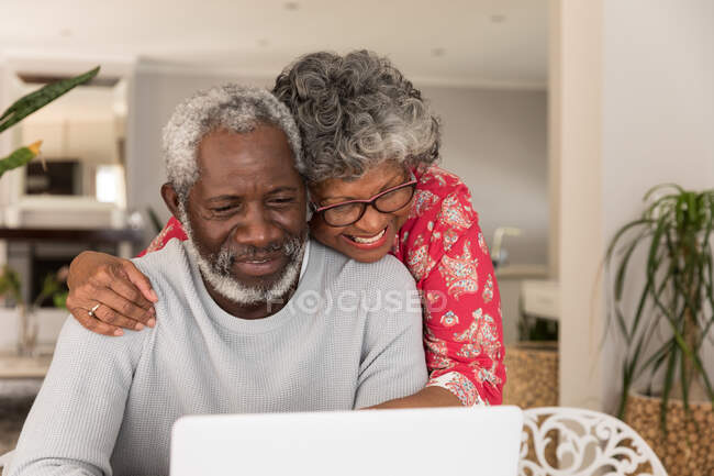 Una coppia afroamericana anziana che trascorre del tempo a casa insieme, distanziamento sociale e isolamento in quarantena durante l'epidemia di coronavirus covid 19, seduta a un tavolo, usando un computer portatile, abbracciando e sorridendo — Foto stock