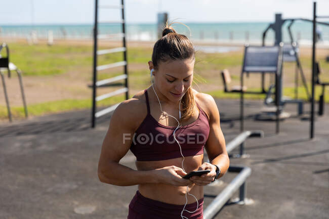 Front view of a sporty Caucasian woman with long dark hair exercising in an outdoor gym during daytime, using her phone with her headphones on. — Stock Photo