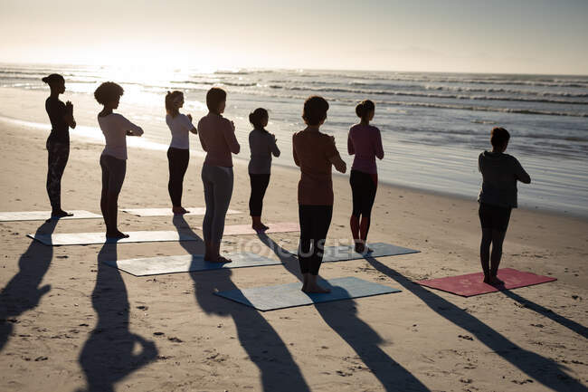Visão traseira de um grupo multi-étnico de amigas que gostam de se exercitar em uma praia em um dia ensolarado, praticando ioga, de pé na posição de ioga. — Fotografia de Stock