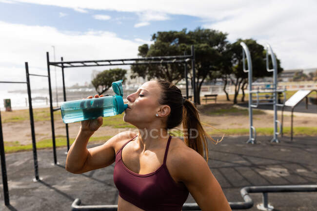 Front view of a sporty Caucasian woman with long dark hair exercising in an outdoor gym during daytime, drinking water from a bottle. — Stock Photo