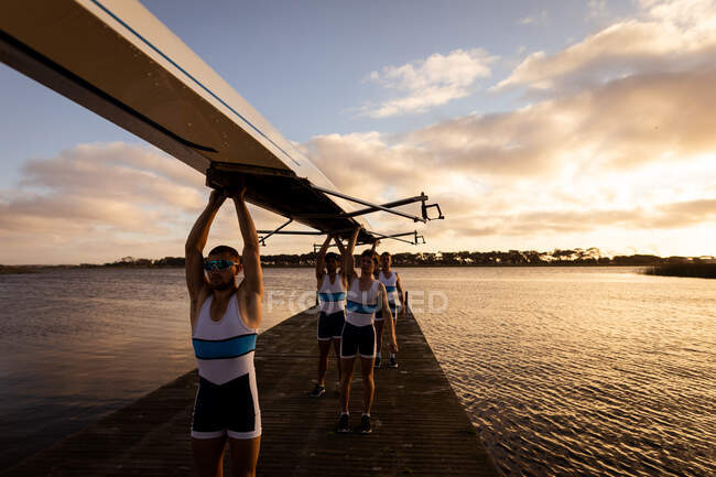 Front view of a rowing team of four Caucasian men carrying a boat above their heads with arms raised, walking along a jetty on the river — Stock Photo