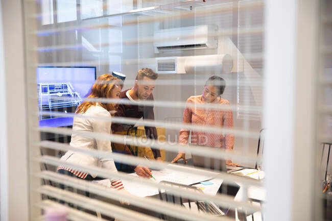 Белая женщина, мужчина и деловая женщина смешанной расы, работающая вместе в современном офисе, используя ноутбук и разговаривая в конференц-зале, через стеклянную стену — стоковое фото