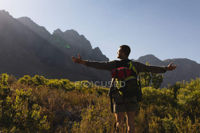 Vista trasera de un hombre caucásico pasándolo bien en un viaje a las montañas, de pie en un campo debajo de las montañas, disfrutando de su vista, sosteniendo sus brazos abiertos, en un día soleado - foto de stock