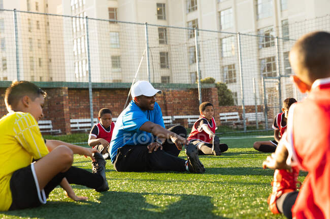 Seitenansicht eines gemischten männlichen Fußballtrainers, der während eines Fußballtrainings auf einem Spielfeld in der Sonne sitzt und einer multiethnischen Gruppe von Jungen-Fußballern Stretchübungen gibt. — Stockfoto