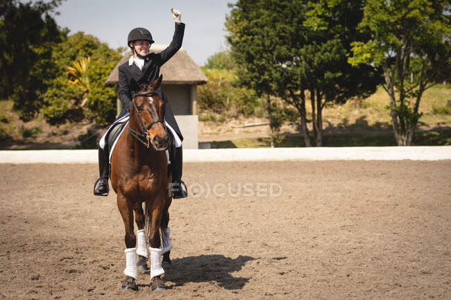 Вид спереди на ловко одетую кавказскую всадницу, сидящую на каштановом коне, машущую рукой и улыбающуюся во время выездного шоу в солнечный день. — стоковое фото