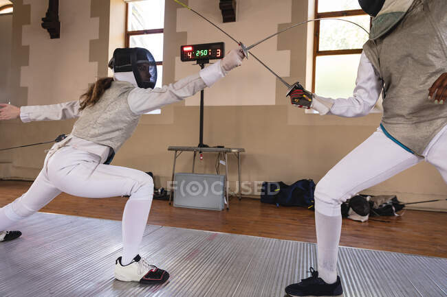 Caucasian and African American sportswomen wearing protective fencing outfits during a fencing training session, taking aim and lunging with their epees. Fencers training at gym. — Stock Photo