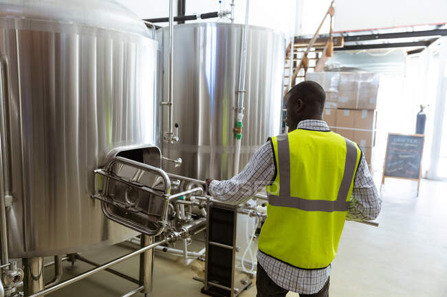 Вид сзади на афроамериканца в клетчатой рубашке и высоко видимом жилете, работающего в пивоварне, проверяющего оборудование. — стоковое фото