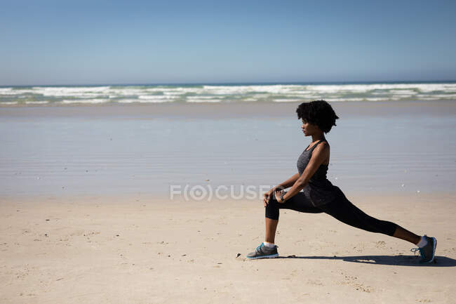 Seitenansicht einer attraktiven afroamerikanischen Frau, die Sportkleidung trägt, Yoga praktiziert, sich in Yogaposition dehnt, am sonnigen Strand. — Stockfoto