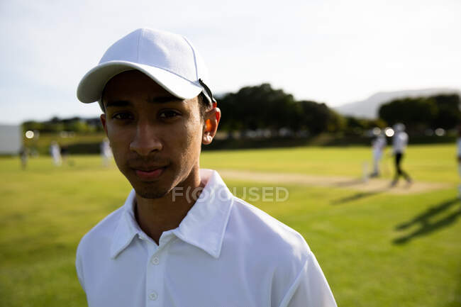 Ritratto di un adolescente asiatico sicuro di sé giocatore di cricket che indossa bianchi di cricket e un cappello, in piedi su un campo da cricket in una giornata di sole guardando alla fotocamera, con altri giocatori che giocano sullo sfondo. — Foto stock