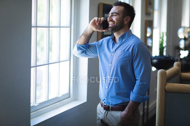 Un homme d'affaires caucasien aux cheveux courts, vêtu d'une chemise bleue, travaillant dans un bureau moderne, debout à la fenêtre et parlant sur son smartphone — Photo de stock