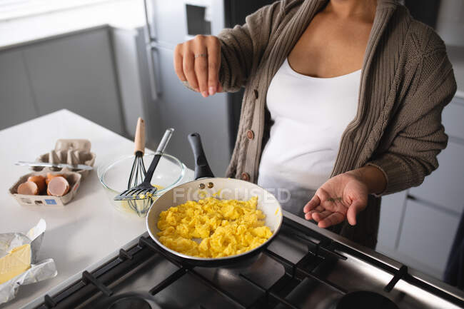 Vista frontal sección media de la mujer relajándose en casa, preparando el desayuno en la cocina, rociando condimentos en huevos revueltos cocinando en una sartén en la encimera - foto de stock