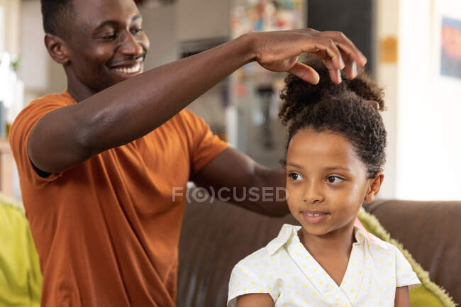 Hombre afroamericano vistiendo una camiseta naranja, distanciamiento social en casa durante el cierre de cuarentena, haciendo una cola de caballo a su hija vistiendo una camisa blanca. - foto de stock