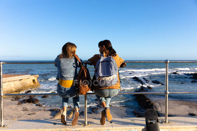 Rückansicht einer kaukasischen und einer gemischten Rasse Mädchen genießen die Zeit zusammen hängen an einem sonnigen Tag, sitzen auf einem Zaun in einer Promenade am Meer, mit Rucksäcken. — Stockfoto