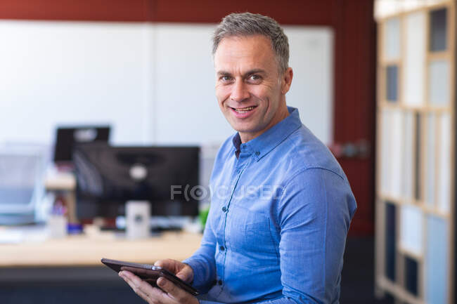 Retrato de un hombre de negocios caucásico con una camisa azul, de pie y sonriente, trabajando en una oficina moderna, mirando a la cámara y usando su tableta - foto de stock