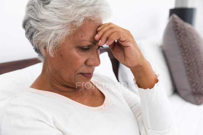 Nahaufnahme einer älteren afroamerikanischen Rentnerin zu Hause, die mit Kopfschmerzen im Bett in ihrem Schlafzimmer sitzt, ihren Kopf berührt und nach unten schaut, sich während der Coronavirus-Pandemie selbst isoliert 19 — Stockfoto