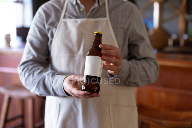 Mitte Barmann, der in einer Mikrobrauerei-Kneipe arbeitet, weiße Schürze trägt und eine Flasche Bier vor sich hält. — Stockfoto