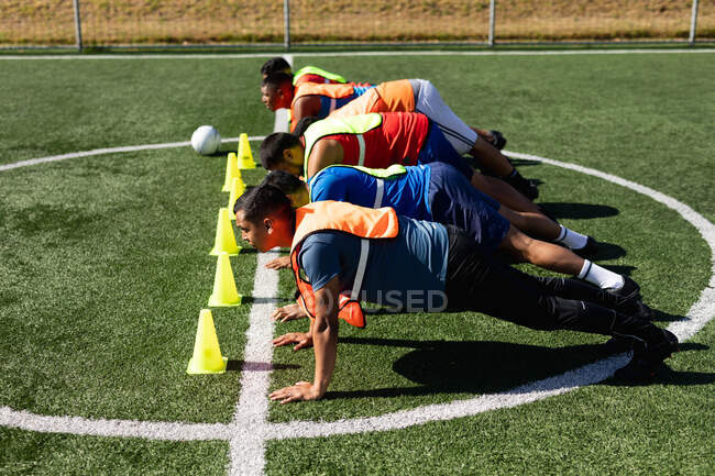 Multi-ethnische Gruppe von männlichen Fußballspielern in Sportkleidung und Weste, die auf einem Sportplatz in der Sonne trainieren und sich mit Ball und Kegel nebeneinander beim Liegestütz aufwärmen. — Stockfoto