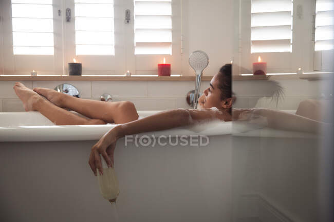 Mixed race woman spending time at home self isolating and social distancing in quarantine lockdown during coronavirus covid 19 epidemic, lying in bathtub relaxing holding champagne glass in bathroom. — Stock Photo