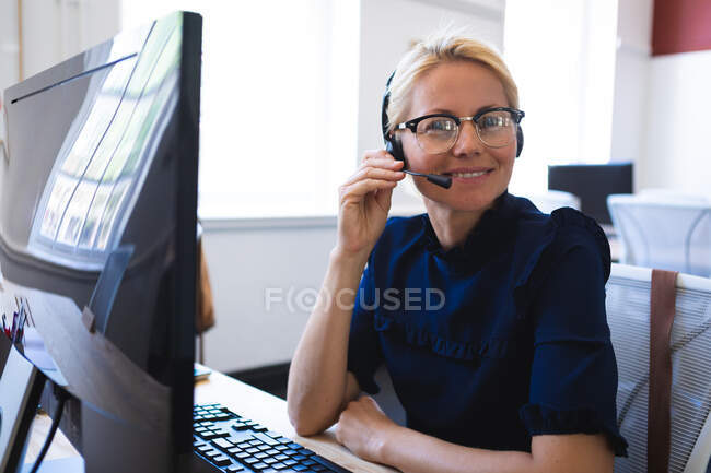 Porträt einer kaukasischen Geschäftsfrau mit kurzen blonden Haaren, die in einem modernen Büro arbeitet, am Schreibtisch sitzt, Headset trägt und in die Kamera blickt — Stockfoto