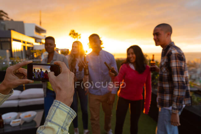 Над плечовим видом багатоетнічної групи друзів, що висять на терасі на даху з небом заходу сонця, тримаючи пляшки пива, один з яких фотографував — стокове фото