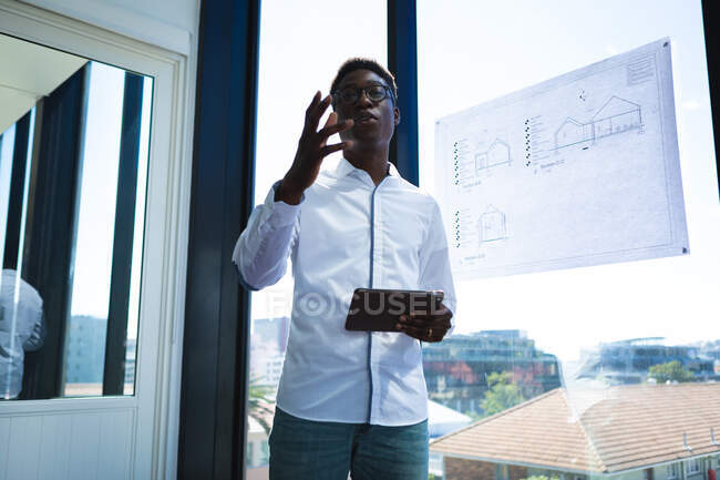 An African American businessman, wearing a white shirt, working in a modern office, standing by the window, holding his tablet and talking, with a plan on the window — Stock Photo