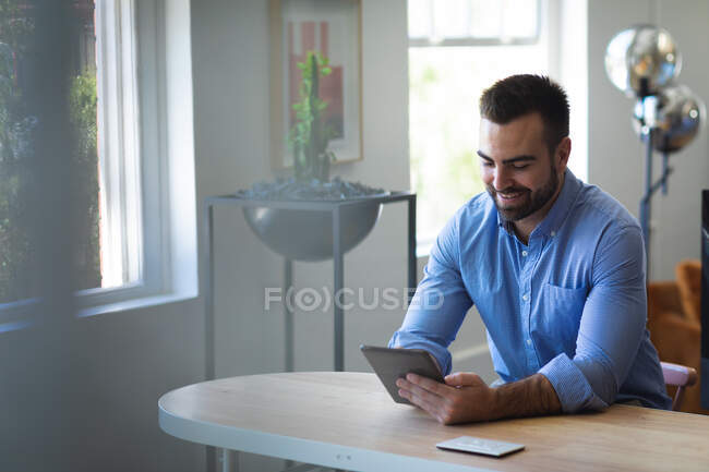 Un hombre de negocios caucásico con el pelo corto, con una camisa azul, trabajando en una oficina moderna, sentado en una mesa y usando su tableta, sonriendo - foto de stock