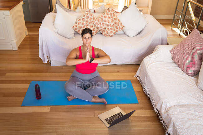 Mulher branca vlogger em casa em sua sala de estar, praticando ioga e usando seu computador portátil. Distanciamento social e auto-isolamento em quarentena . — Fotografia de Stock