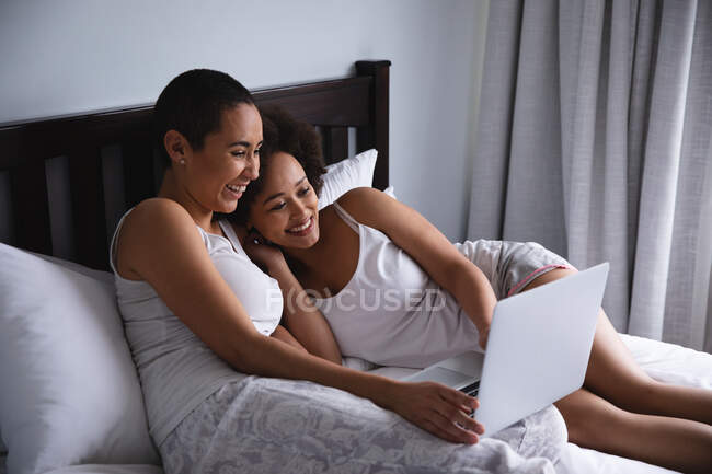 Vista laterale di una coppia mista di donne che si rilassano a casa in camera da letto, si siedono sul letto usando un computer portatile insieme e sorridono — Foto stock