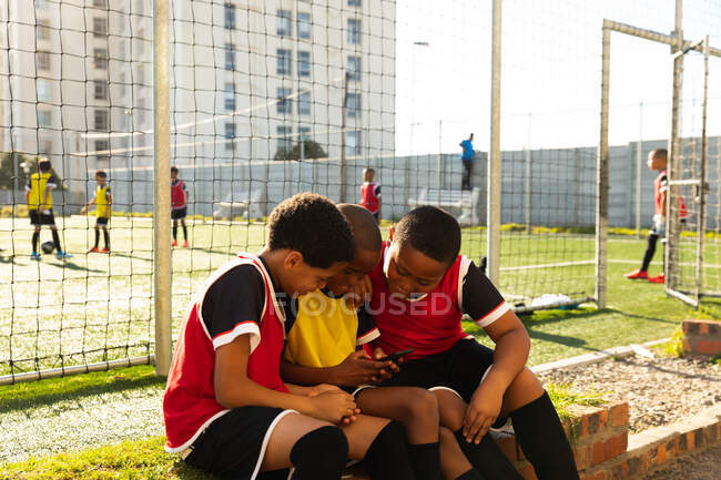 Вид збоку на багатоетнічну групу хлопчиків-футболістів, які носять командну смугу, сидячи на ігровому полі, тримаючи і використовуючи смартфон разом на сонці — стокове фото