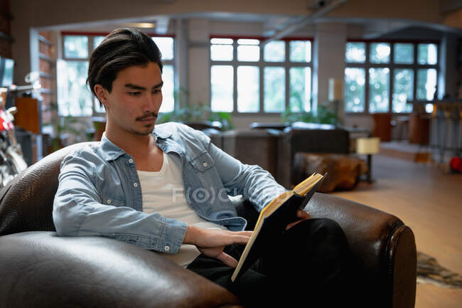 Side view of a young mixed race man enjoying time at home, resting in his sitting room, sitting in the armchair, reading a book. — Stock Photo
