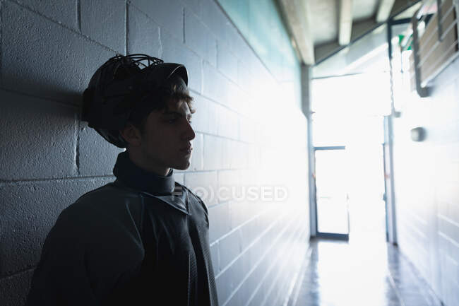 Vista laterale di un adolescente caucasico giocatore di hockey su prato maschile, in piedi da solo in un corridoio indossando un casco, preparando e concentrandosi prima di una partita — Foto stock