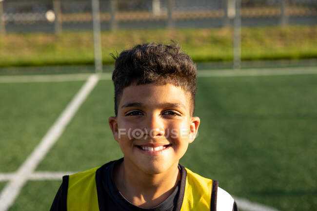 Retrato de cerca de un jugador de fútbol de raza mixta confiado usando una tira de equipo, de pie en un campo de juego en el sol, mirando a la cámara y sonriendo - foto de stock