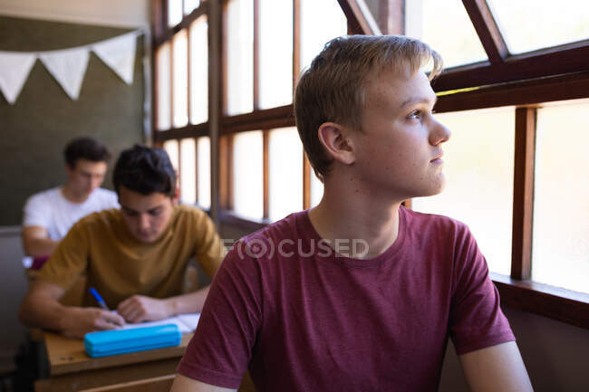 Front view close up of a Caucasian teenage boy sitting at a desk in a school classroom looking out of the window, with classmates sitting at desks working in the background — Stock Photo