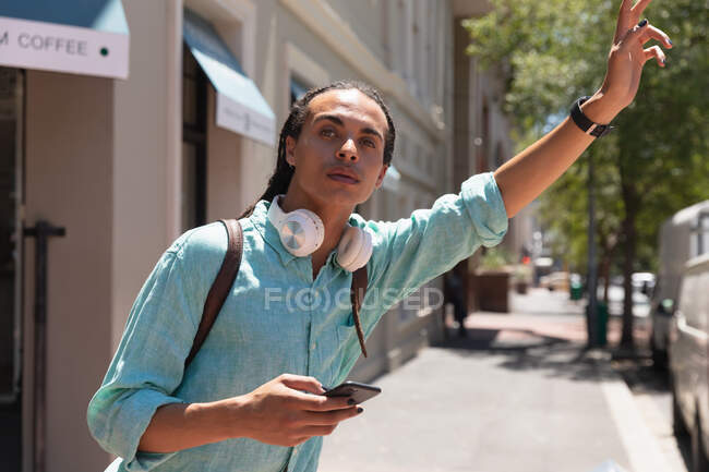 Vista frontal de un hombre de raza mixta con largas rastas en la ciudad en un día soleado, parado en la calle, usando un teléfono inteligente y levantando la mano para detener un taxi. - foto de stock