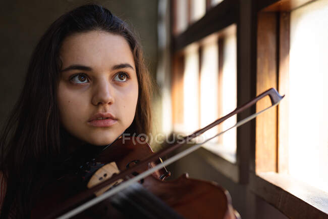 Retrato de cerca de una adolescente músico caucásico sentado junto a una ventana tocando el violín en una escuela secundaria - foto de stock