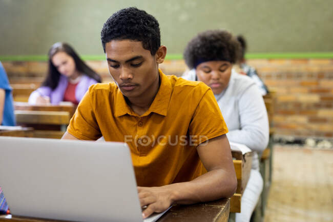 Vista frontal de un adolescente de raza mixta en un aula de la escuela sentado en el escritorio, concentrándose y utilizando la computadora portátil, con compañeros de clase adolescentes masculinos y femeninos sentados en escritorios trabajando en el fondo - foto de stock