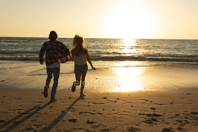 Pareja caucásica corriendo en una playa durante una puesta de sol, tomados de la mano y mirando al mar - foto de stock
