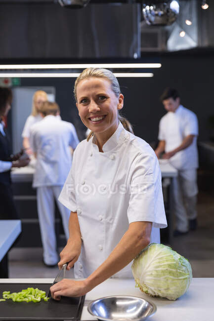 Портрет кавказької жінки-кухарки, що різала овочі, дивлячись на камеру і посміхаючись, з іншими шеф-кухарями на задньому плані. Клас куховарства на кухні.. — стокове фото