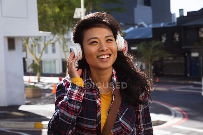 Front view of a mixed race woman with long dark hair out and about in the city streets during the day, wearing headphones, a checked shirt and walking in a city street with buildings in the background. — Stock Photo