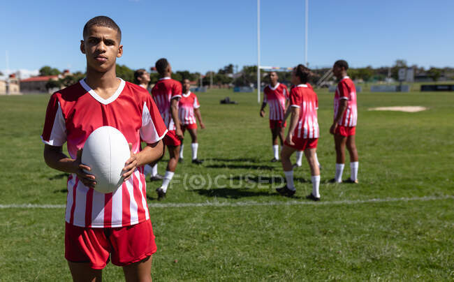 Front view of a teenage mixed race male rugby player wearing red and white team strip, standing on a playing field, holding a rugby ball and looking at camera with other players warming up in the background — Stock Photo