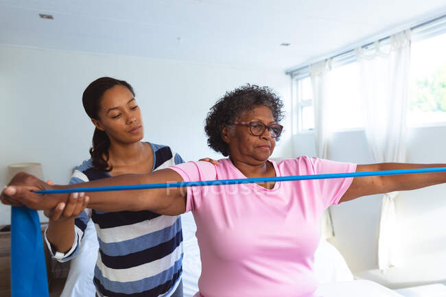 Senior Mixed Race Frau, die Zeit zu Hause verbringt und von einer Mixed Race Krankenschwester besucht wird, die ihre Arme mit Gummiband ausspannt — Stockfoto