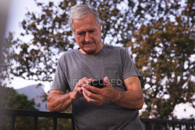Красивый пожилой кавказский мужчина, наслаждающийся пенсией, в саду на солнце, переписывающийся с мобильным телефоном, самоизолирующийся во время пандемии коронавируса covid19 — стоковое фото