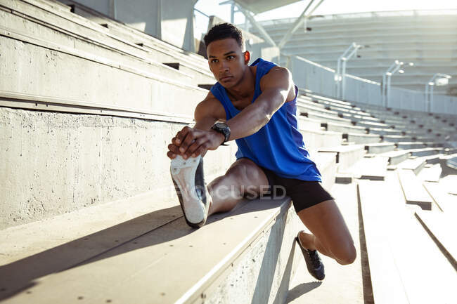 Vista frontal de un atleta masculino de raza mixta practicando en un estadio deportivo, sentado en las gradas y estirándose, tirando de su pie - foto de stock